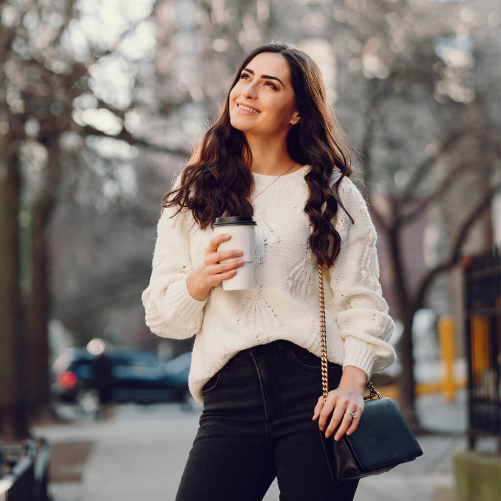 Femme souriante avec un café à emporter, portant un pull en tricot blanc et un sac à main noir.