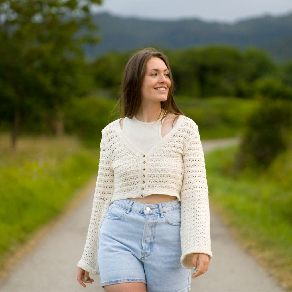 Femme souriante portant un cardigan en tricot crémeux et un short en jean, marchant sur un chemin dans la nature.