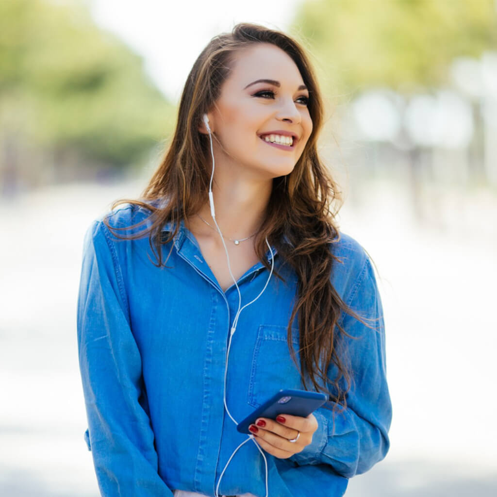 jeune femme souriante portant une chemise en jean, écoutant de la musique avec des écouteurs et un téléphone dans la main