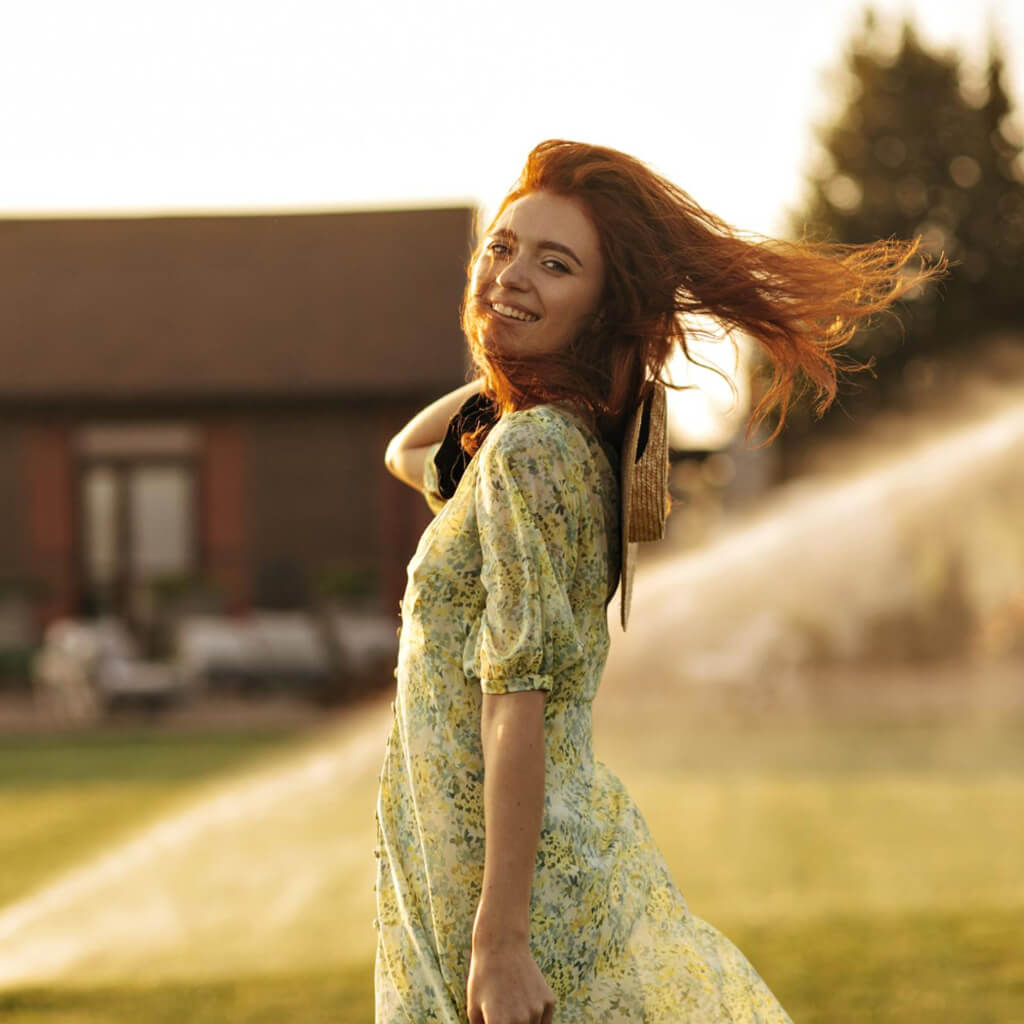Femme souriante en robe jaune dans un jardin, cheveux au vent, illuminée par le soleil du soir.