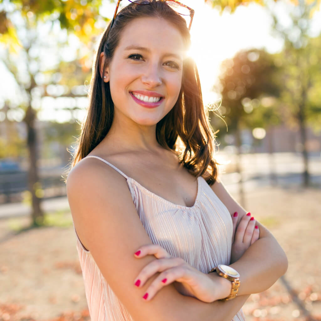 Femme souriante en été avec rouge à lèvres aux plantes et lunettes de soleil, devant des arbres.