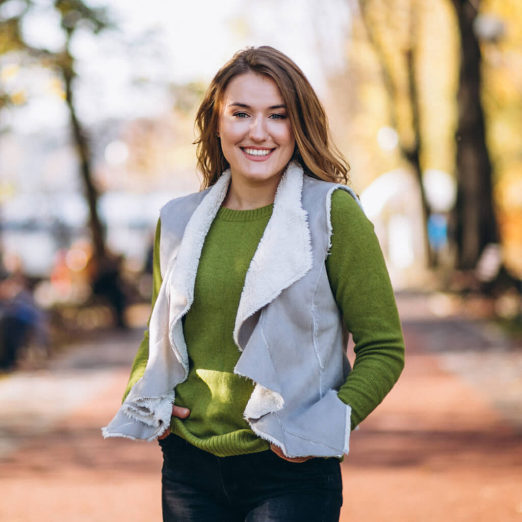 Femme souriante portant un gilet gris et un pull vert dans un parc en automne.