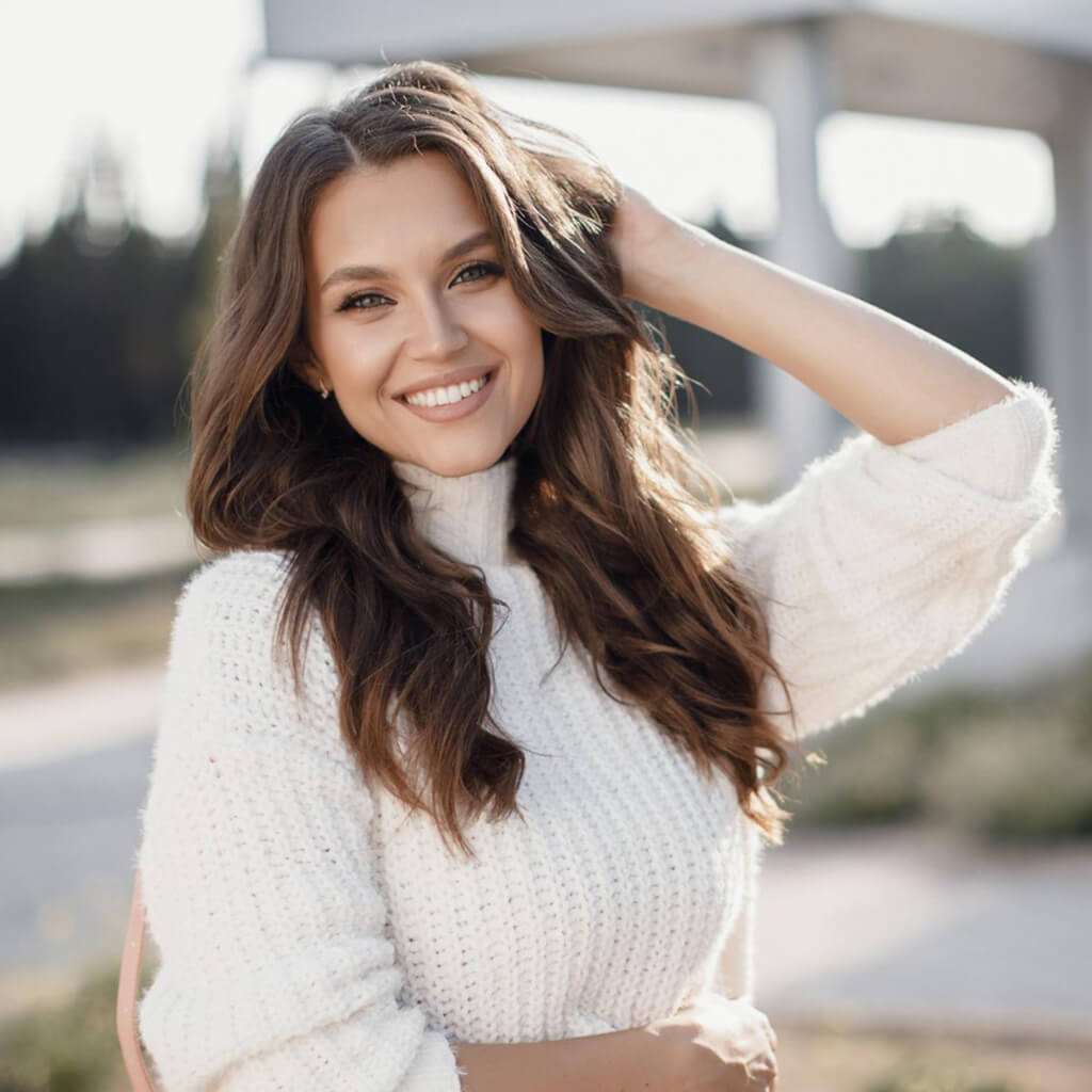 Femme souriante portant un pull en laine, avec des cheveux ondulés, dans un cadre en plein air.