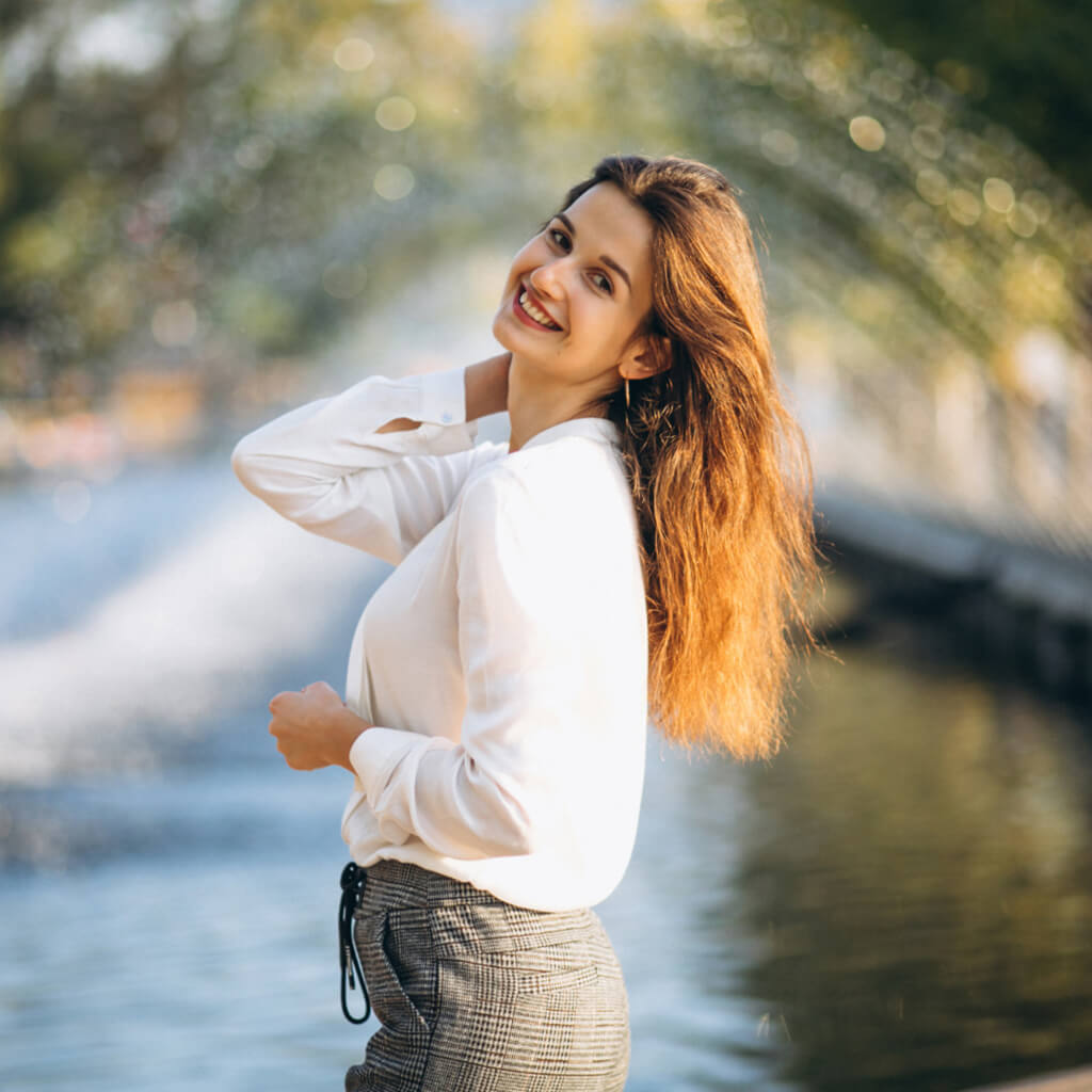 Femme souriante avec cheveux longs, portant une chemise blanche près d'une fontaine.