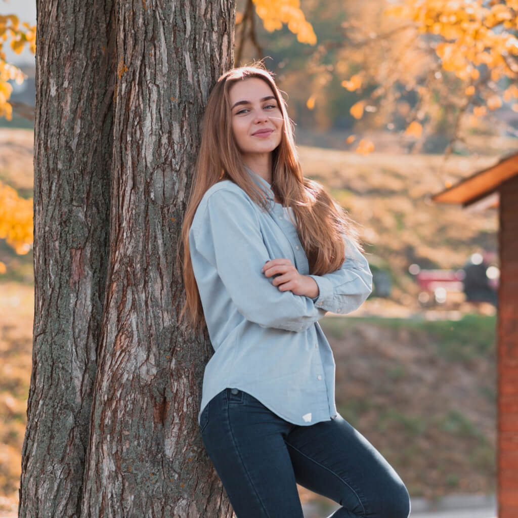 Jeune femme souriante posant en jeans et chemise près d'un arbre pendant l'automne.