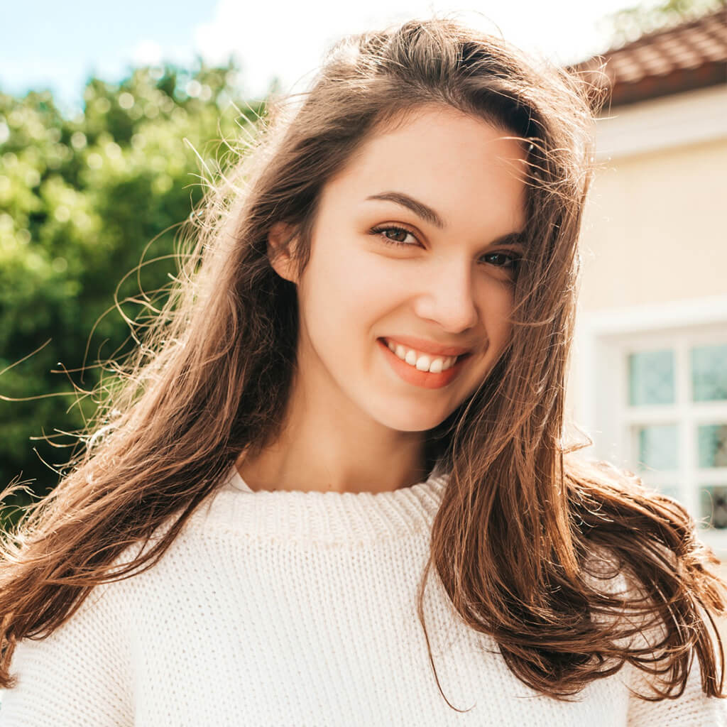 Femme souriante avec des cheveux longs, portant un pull blanc, prenant un moment pour sourire en extérieur.