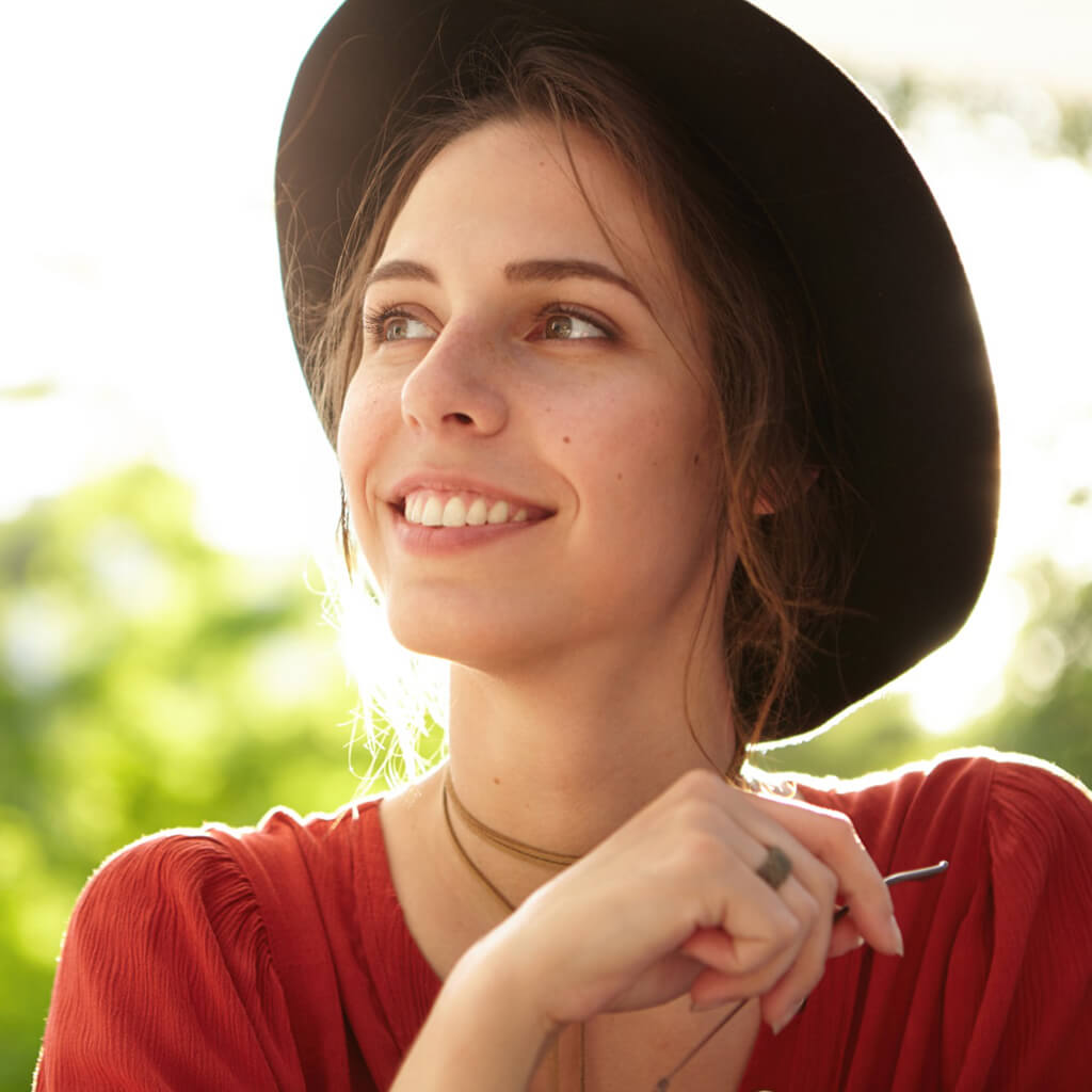 Jeune femme souriante portant un chapeau noir, avec une ambiance naturelle et lumineuse en arrière-plan.