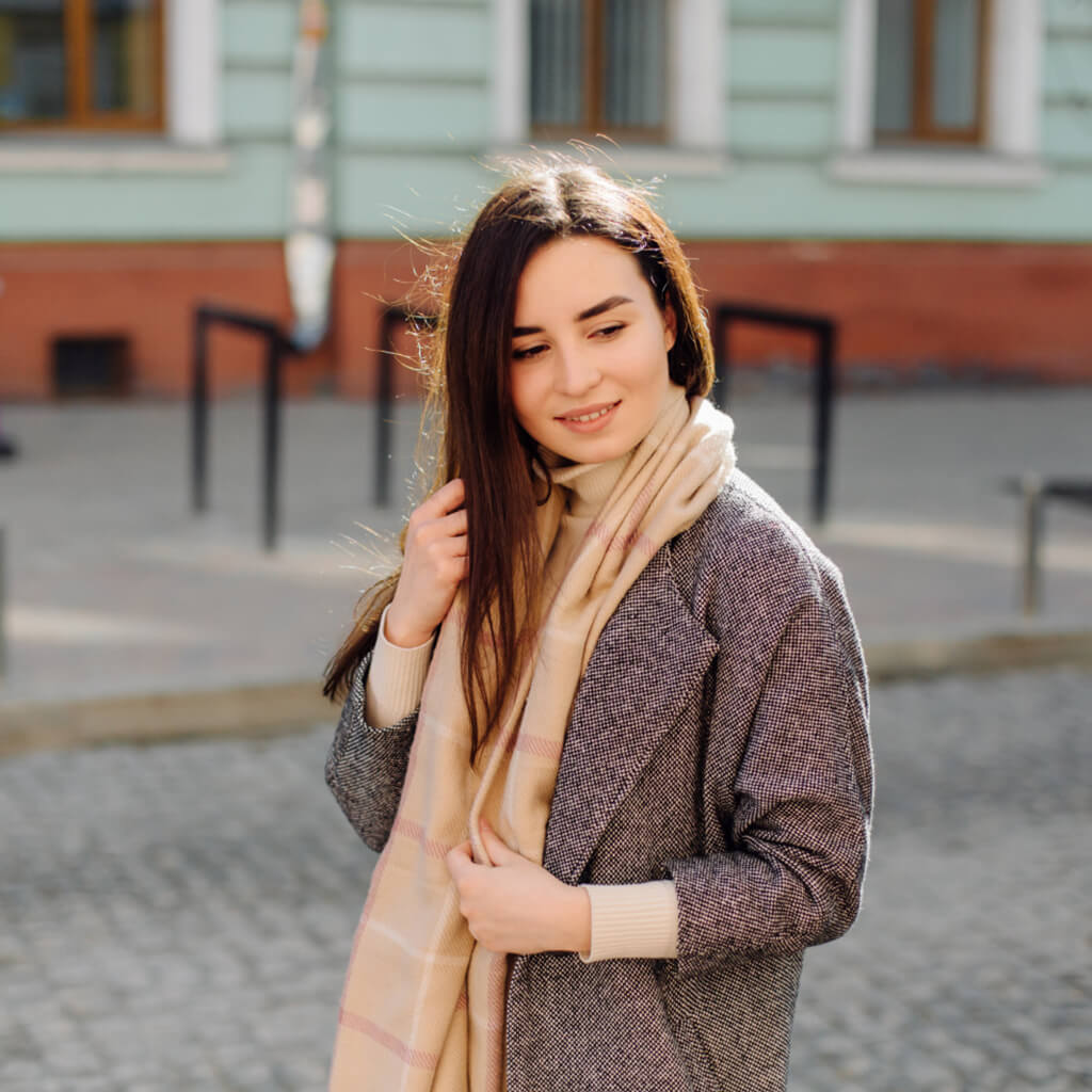 Femme souriante avec écharpe élégante, portant un manteau sophistiqué dans une rue ensoleillée.